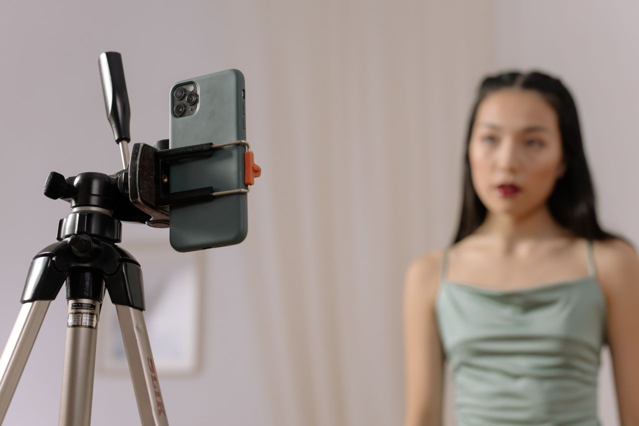 A woman is standing in front of a smartphone mounted on a tripod, taking part in indoor vlogging content.